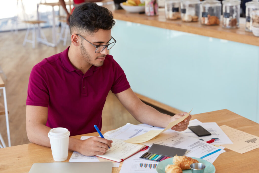 “A young man wearing glasses and a maroon polo shirt sits at a café table, reviewing documents and writing notes in a notebook. A coffee cup, smartphone, charts, and pastries are placed on the table around him.”