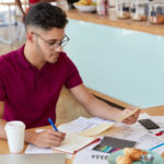 “A young man wearing glasses and a maroon polo shirt sits at a café table, reviewing documents and writing notes in a notebook. A coffee cup, smartphone, charts, and pastries are placed on the table around him.”